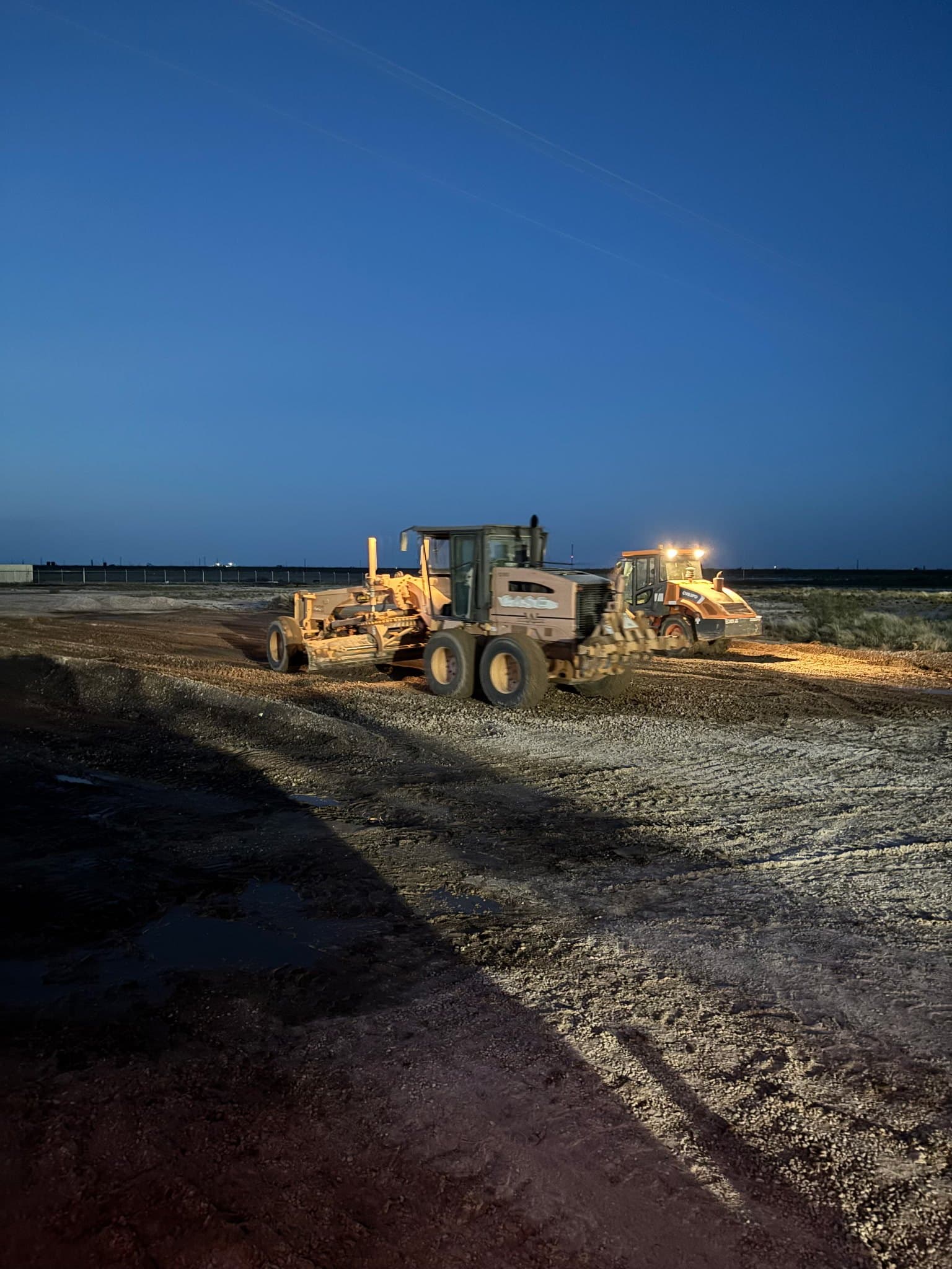 Grader holding grade alongside a roller on a Permian Basin pad at dusk